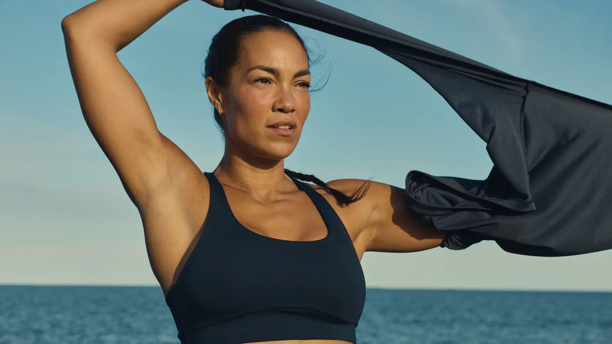 Woman in a dark navy Tripulse TENCEL™ Lyocell sports bra by the sea, pulling a top off over her head after a hot workout.