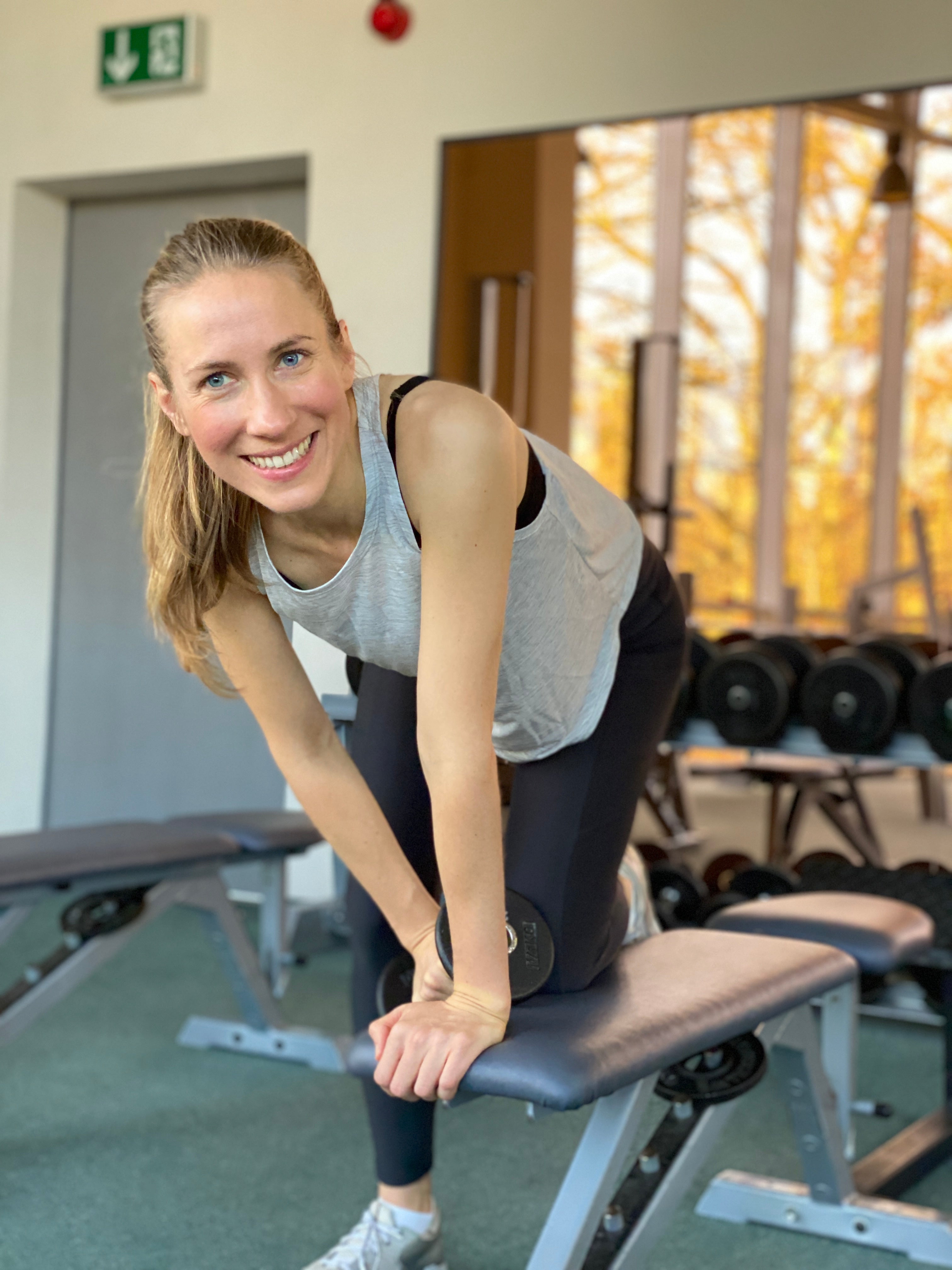Smiling woman leaning on a workout bench in a gym wearing a grey top and leggings.