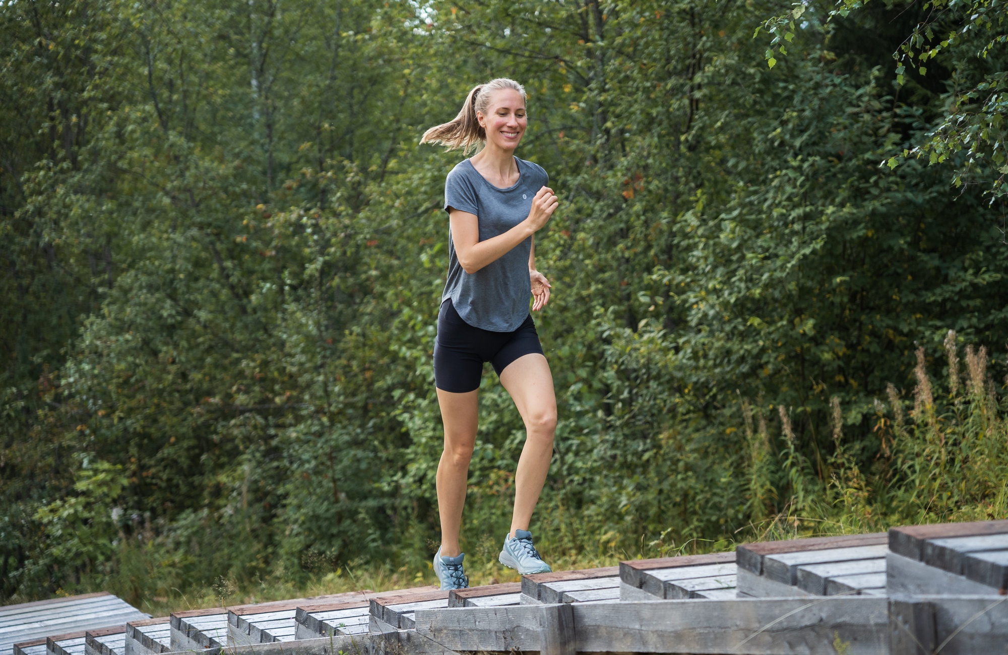 Woman jogging up wooden outdoor steps in a blue Tripulse T-shirt and shorts.