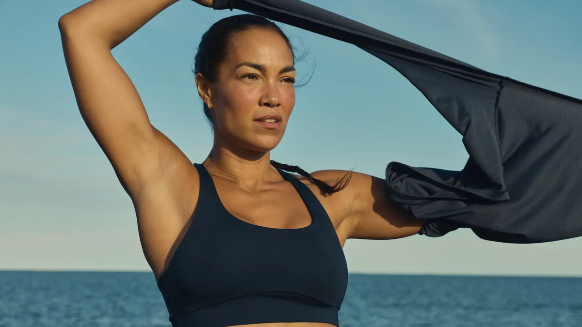 Woman in a dark navy Tripulse TENCEL™ Lyocell sports bra by the sea, pulling a top off over her head after a hot workout.