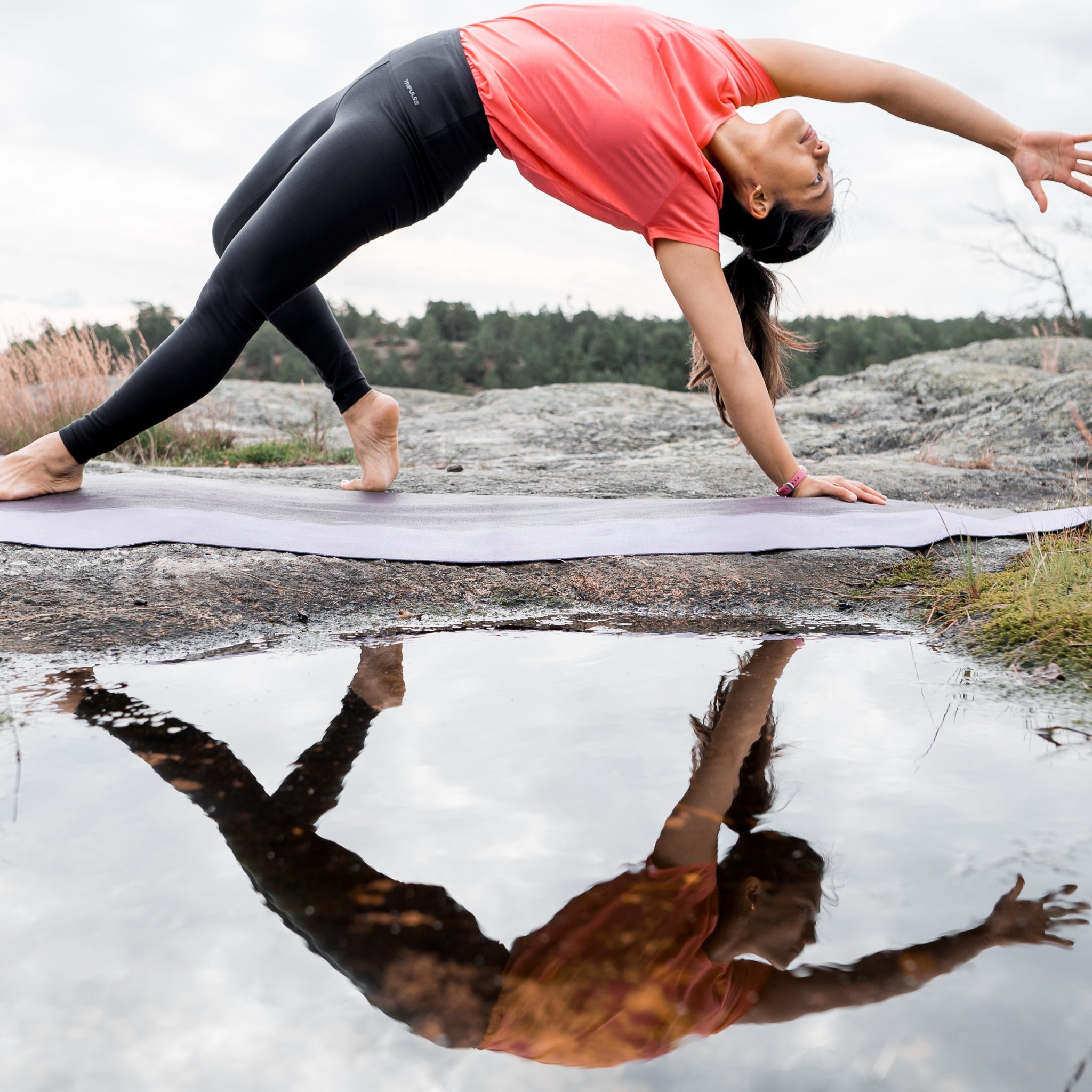 Woman doing a yoga wheel pose on a mat by a lake, reflected in the water.